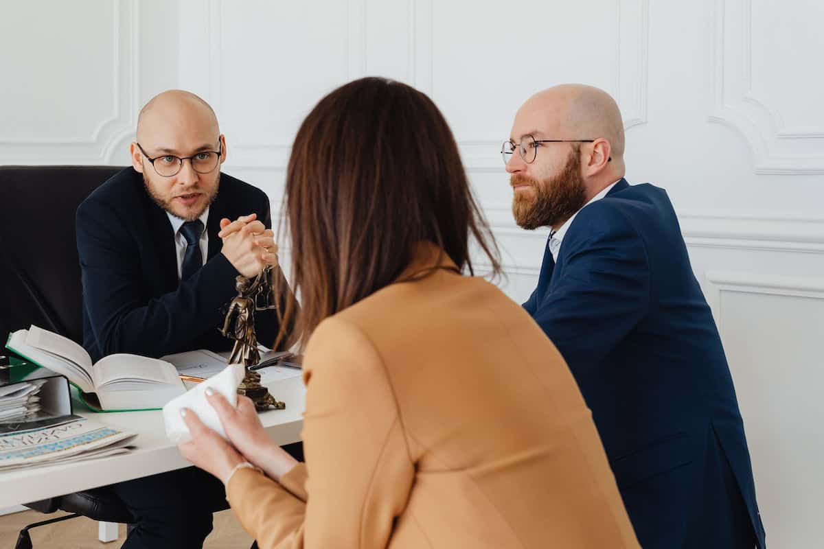 Two suited men and a woman sit at a white desk in a bright office; one man speaks while the other listens as papers and a small statue sit on the desk